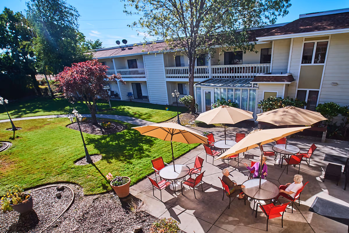 Outdoor courtyard of a senior living facility with round tables, large umbrellas, red chairs, and several seated residents in front of a two-story building.