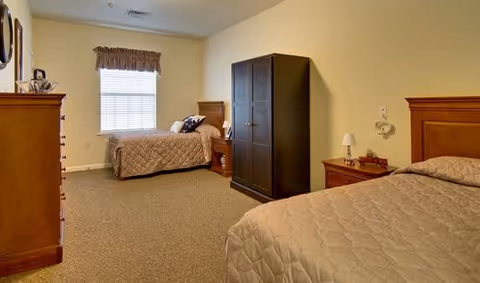 A senior living facility bedroom with two beds, each with beige bedspreads. There is a wooden dresser on the left, a dark wardrobe in the center, and a nightstand with a lamp between the beds. A window with a valance is on the far wall, and the room has beige walls and carpeted floor.