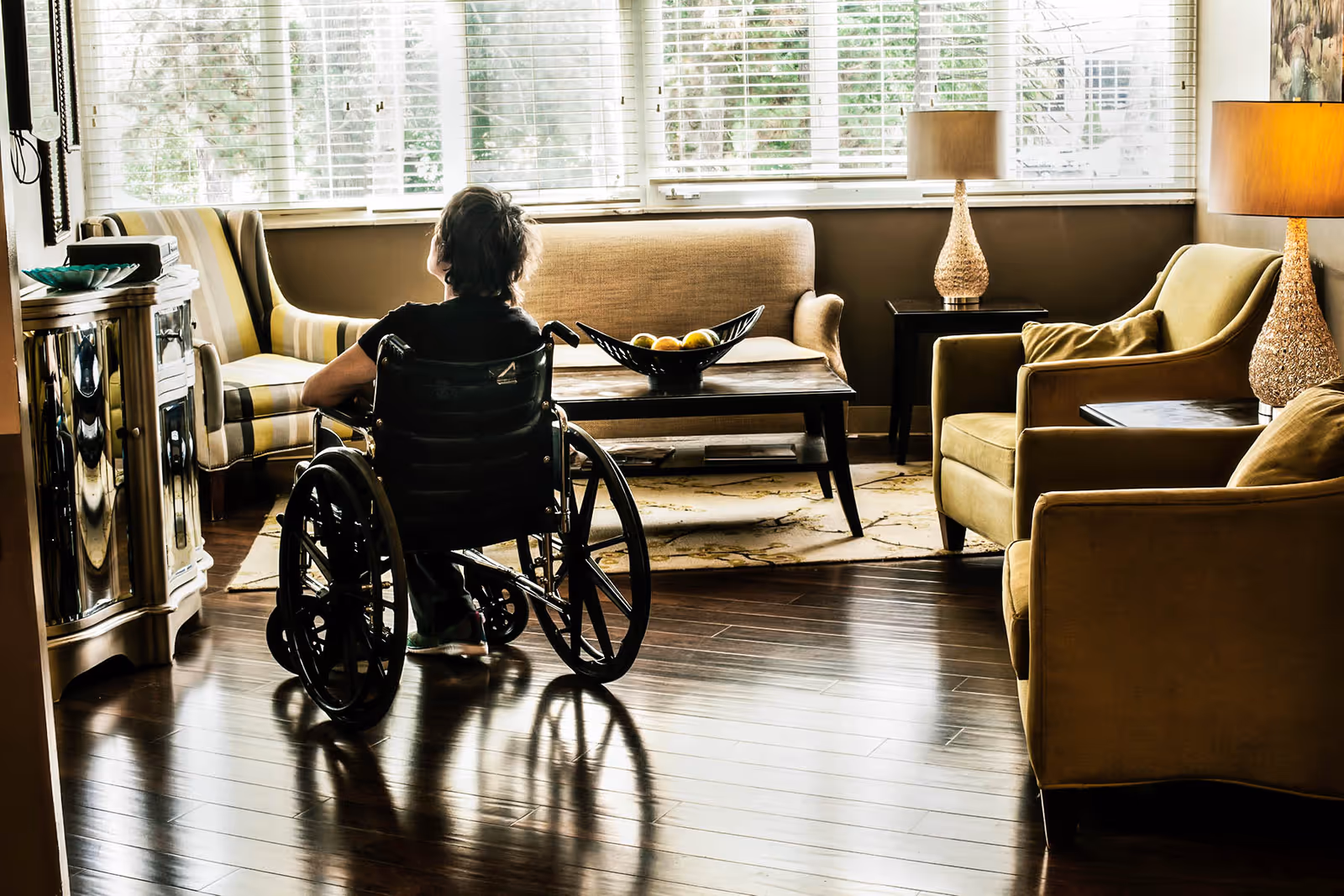 A person in a wheelchair sits in a sunlit living room with armchairs, a coffee table, lamps, and hardwood floors.