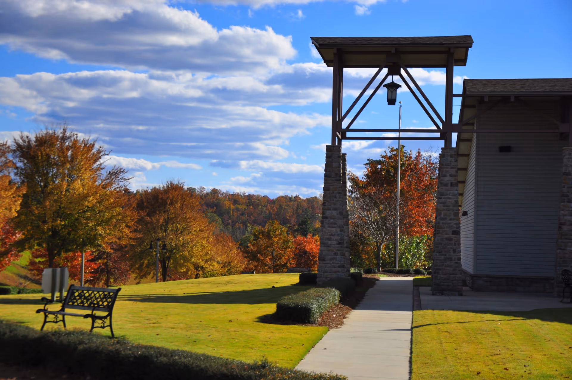 Outdoor view of a senior living facility with a paved walkway leading under a wooden and stone pergola structure. There is a bench on the left side on a well-maintained grassy lawn with autumn-colored trees in the background under a partly cloudy blue sky.