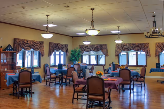 A spacious dining room with multiple tables covered in blue and purple tablecloths, each set with chairs and small flower arrangements. The room has large windows with decorative valances, wooden flooring, and ceiling lights providing warm illumination.