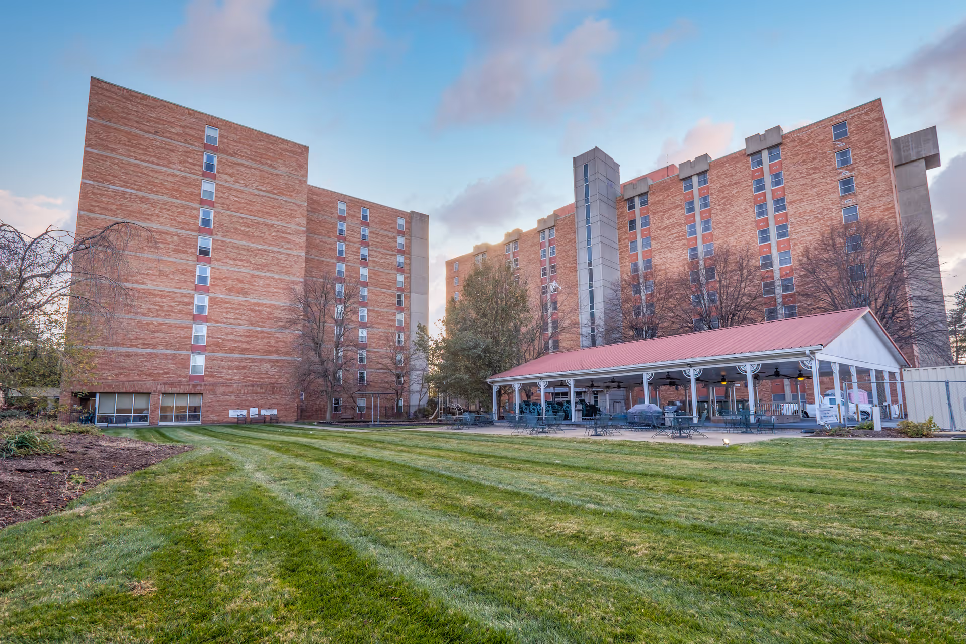 Exterior view of Chapel House Louisville showing two large multi-story brick buildings with many windows, a covered outdoor seating area with tables and chairs, and a well-maintained grassy lawn in the foreground under a partly cloudy sky.