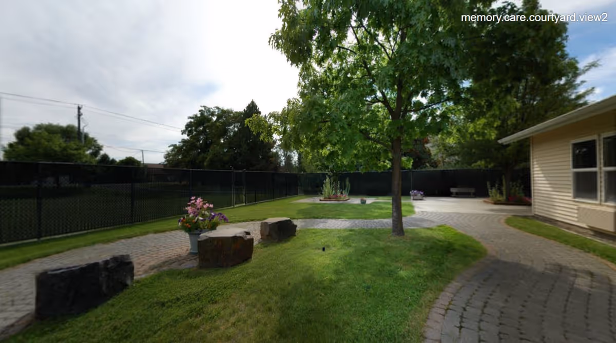Outdoor courtyard area with a paved walkway, green grass, a tree in the center, flower pots with colorful flowers, and a bench near a beige building under a partly cloudy sky.