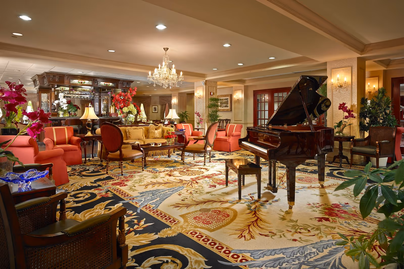 Elegant communal lounge with a grand piano, upholstered seating, chandeliers, and floral carpets.
