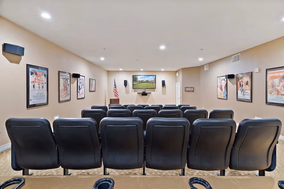 A small movie theater room with three rows of black cushioned seats facing a wall-mounted flat screen TV. The walls are beige and decorated with framed vintage movie posters. There are speakers mounted on the walls and an American flag in the corner near the TV.