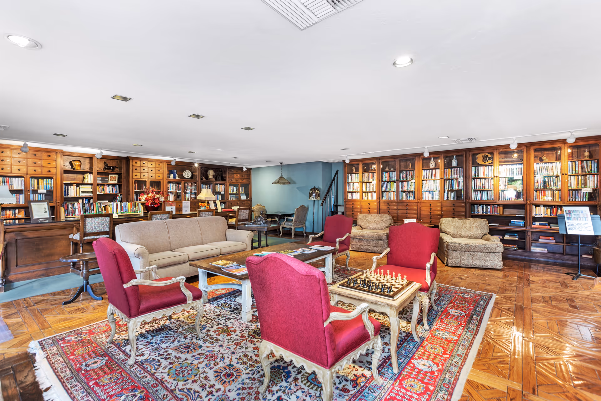 Spacious library-style lounge with bookshelves lining the walls, upholstered sofas and chairs arranged around coffee tables and a chessboard on a decorative rug.