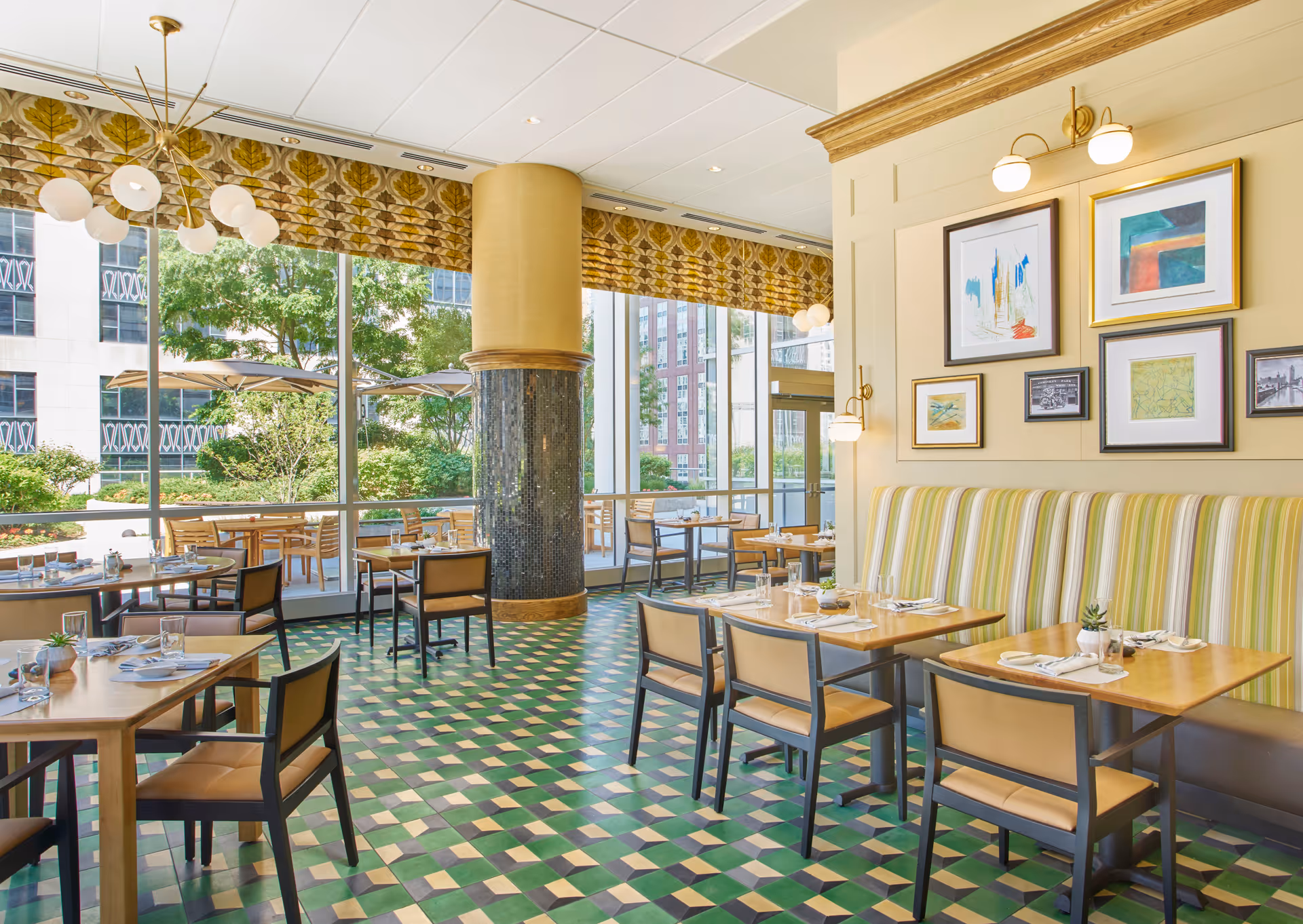 Bright dining room with large windows overlooking an outdoor patio area with tables and umbrellas. The room features wooden tables set with glasses and napkins, cushioned chairs, a striped banquette along one wall, framed artwork, and a patterned green and yellow tiled floor.