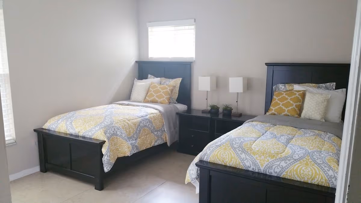 Bright bedroom with two twin beds, matching dark wood headboards and yellow-gray patterned bedding separated by a central nightstand and window.