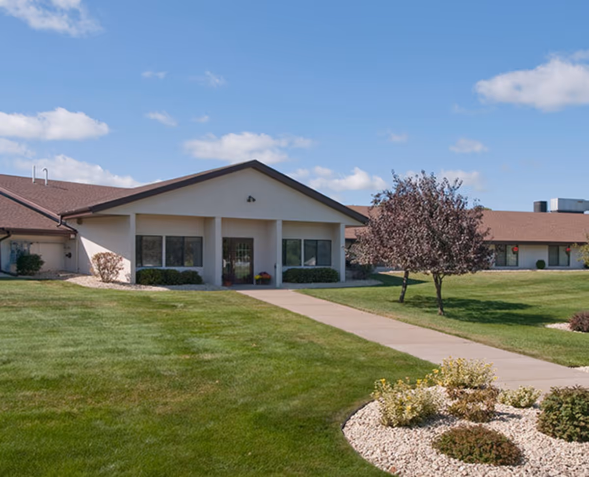 Single-story building with a brown roof and beige walls, surrounded by a well-maintained green lawn, small trees, and landscaped bushes under a blue sky with scattered clouds.
