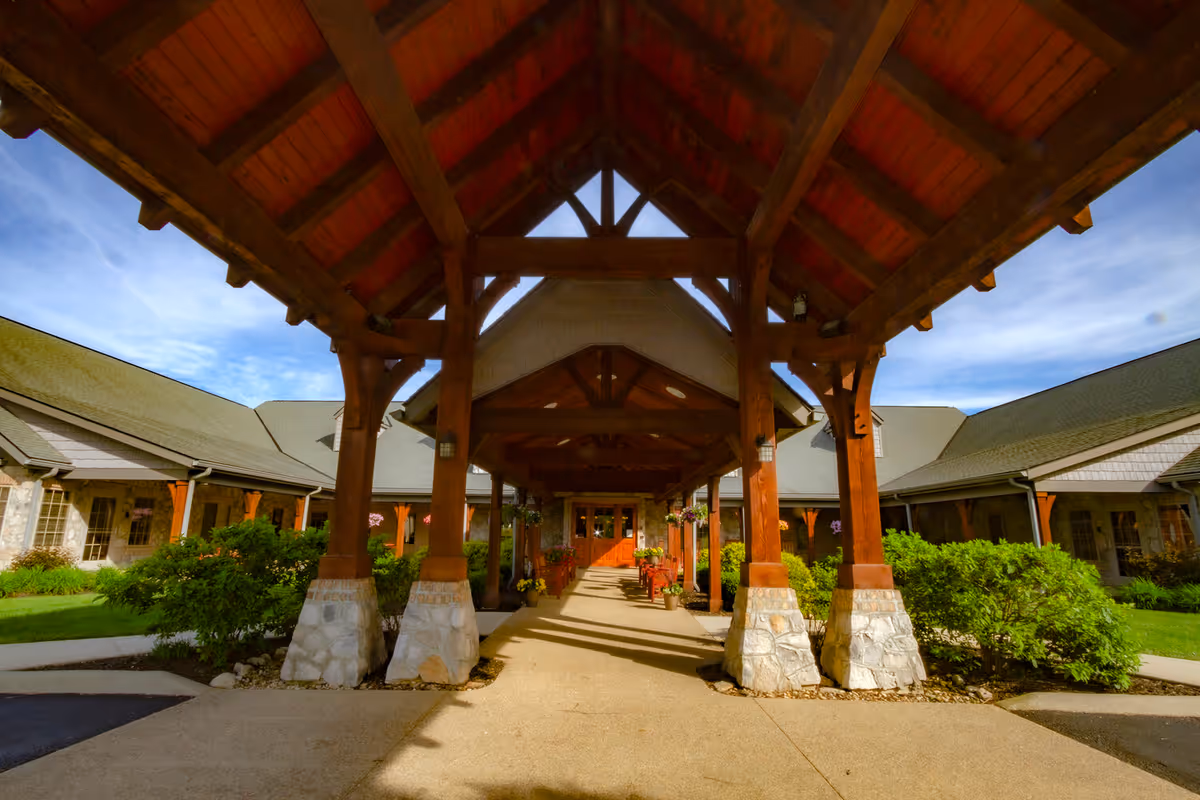 Covered entrance of a building with wooden beams and stone bases, surrounded by greenery and a clear blue sky.