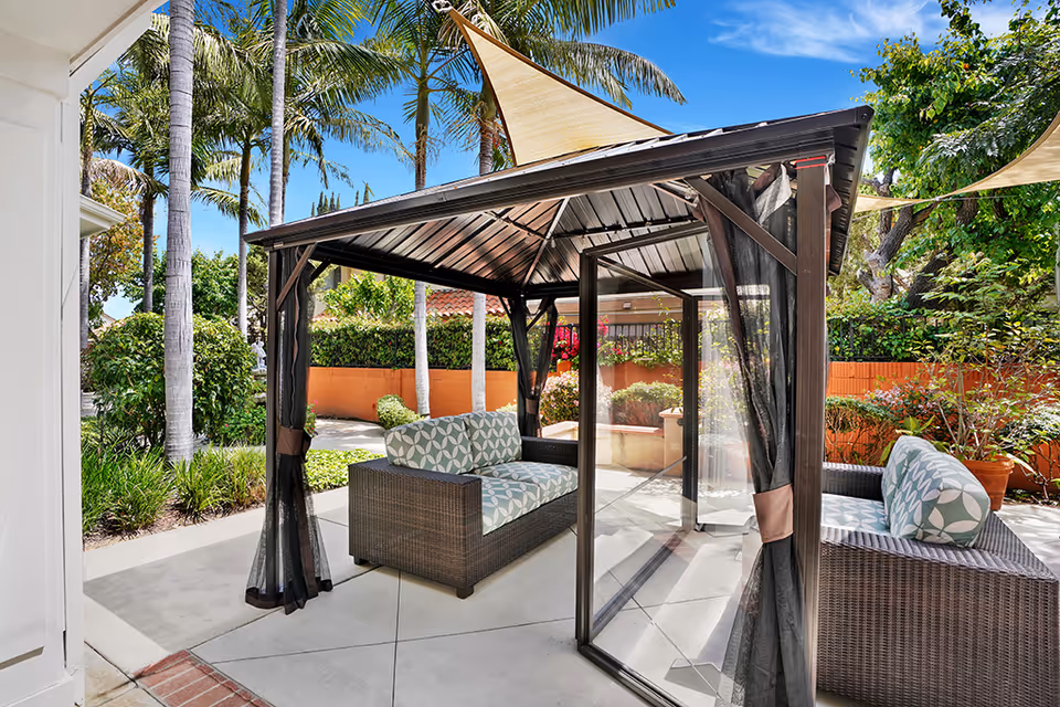 Outdoor seating area with two cushioned wicker sofas under a metal gazebo with a triangular sunshade above. The area is surrounded by palm trees, green shrubs, and a brick wall with potted plants, under a clear blue sky.