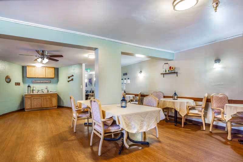 Interior view of a dining room in a senior living facility with several tables covered with beige tablecloths and chairs arranged around them. The room has light green walls, wooden flooring, and ceiling lights. A small cabinet with coffee and tea supplies is visible in the background under a ceiling fan.