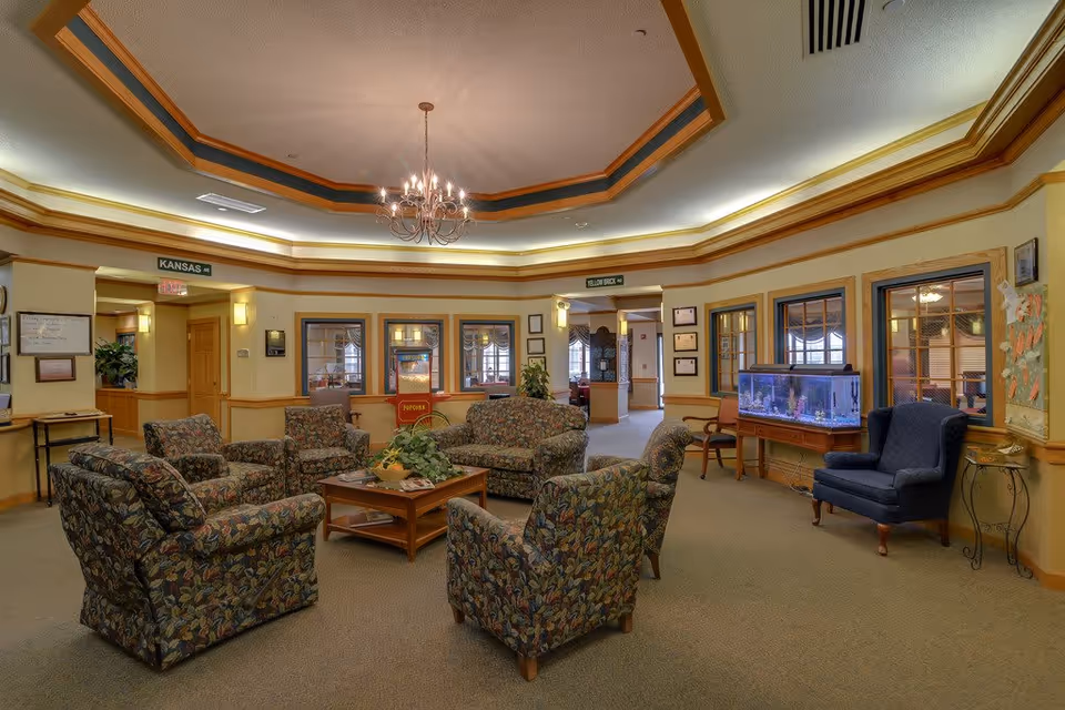 A cozy senior living facility common area with floral upholstered armchairs and a matching sofa arranged around a wooden coffee table with a plant centerpiece. The room features a decorative chandelier hanging from a tray ceiling with recessed lighting. There is a fish tank on a wooden stand against the wall, a blue armchair nearby, and windows with wooden frames. Doorways labeled 'Kansas' and 'Yellow Brick' lead to other rooms.