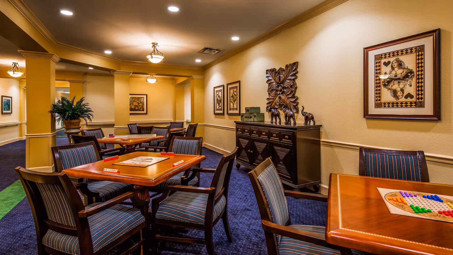 A well-lit interior room with multiple wooden tables and striped cushioned chairs arranged for board games. The tables have game boards and pieces set up. The room features beige walls with framed artwork, decorative sculptures on a sideboard, and recessed ceiling lights.