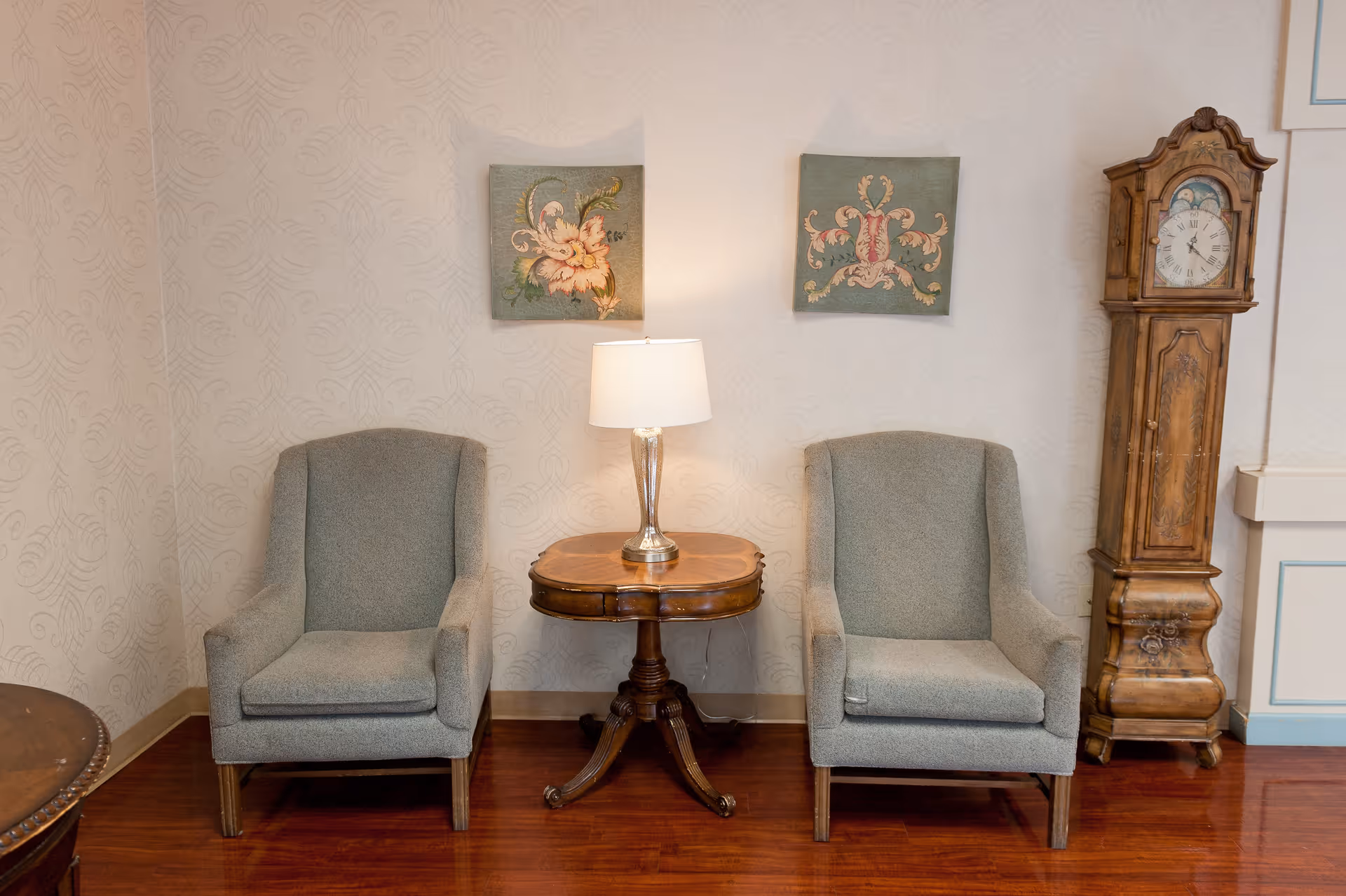 Two gray upholstered armchairs with wooden legs are positioned on either side of a small wooden side table with a lamp on it. Above the chairs are two framed floral artworks. To the right of the chairs is a tall wooden grandfather clock. The room has light-colored patterned wallpaper and wooden flooring.