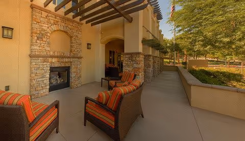 Outdoor patio area with cushioned wicker chairs, a stone fireplace, pergola overhead, and a landscaped walkway.