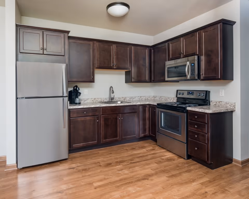 A modern kitchen with dark wood cabinets, a stainless steel refrigerator, a built-in microwave above a black electric stove, a single basin sink with a faucet, and a coffee maker on the countertop. The floor is wood, and the walls are painted white.