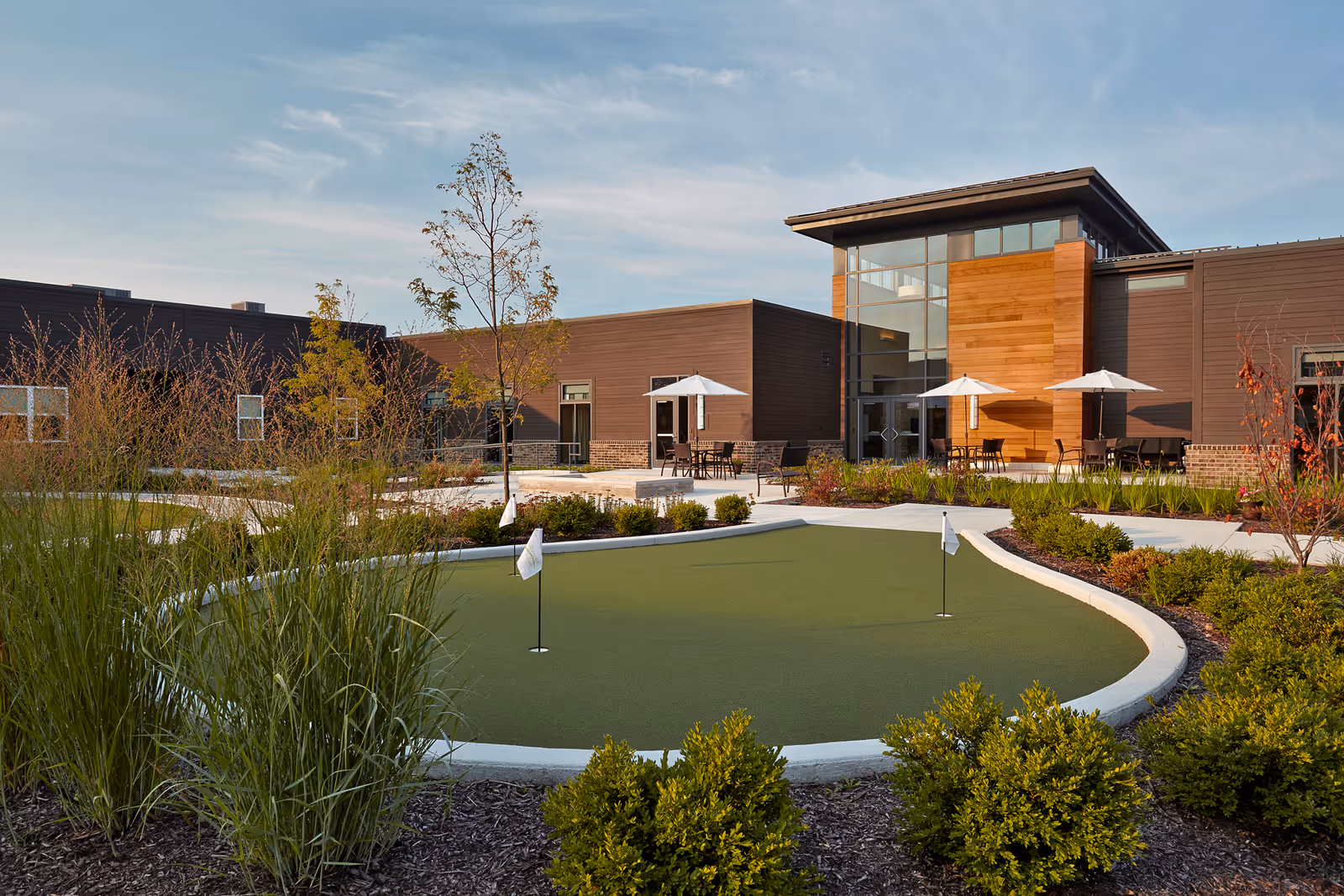 Outdoor courtyard with a small putting green and patio tables with umbrellas in front of a modern senior living building.