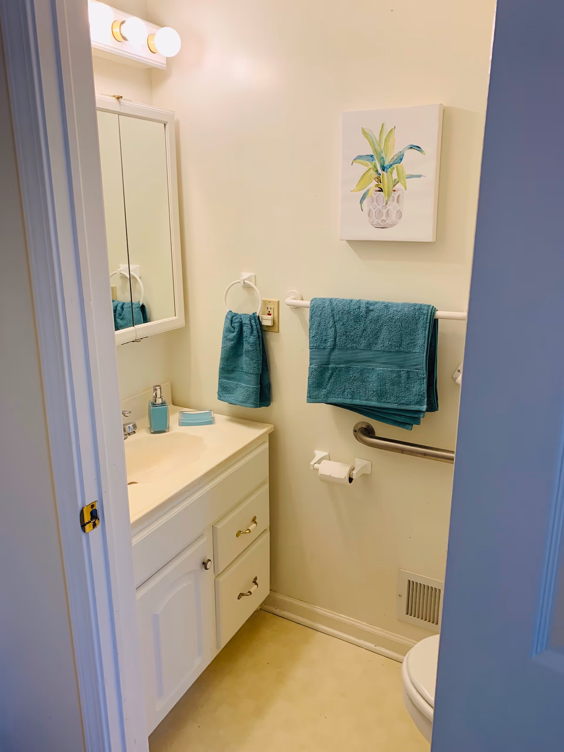 A small bathroom with a white vanity and mirror, teal towels, wall art, a grab bar and a toilet.