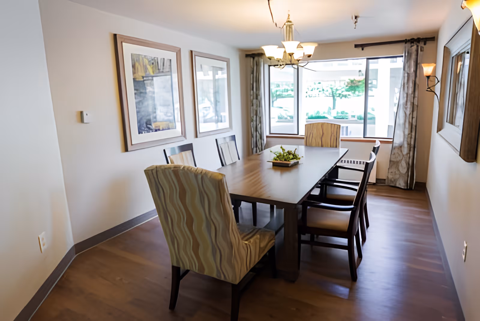 A dining room with a rectangular wooden table surrounded by six chairs, two of which have patterned upholstery. The room has wooden flooring, two framed abstract paintings on the wall, a chandelier overhead, and large windows with curtains letting in natural light.