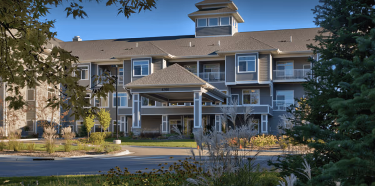Exterior view of a multi-story senior living facility building with a covered entrance, surrounded by landscaped greenery and trees under a clear blue sky.
