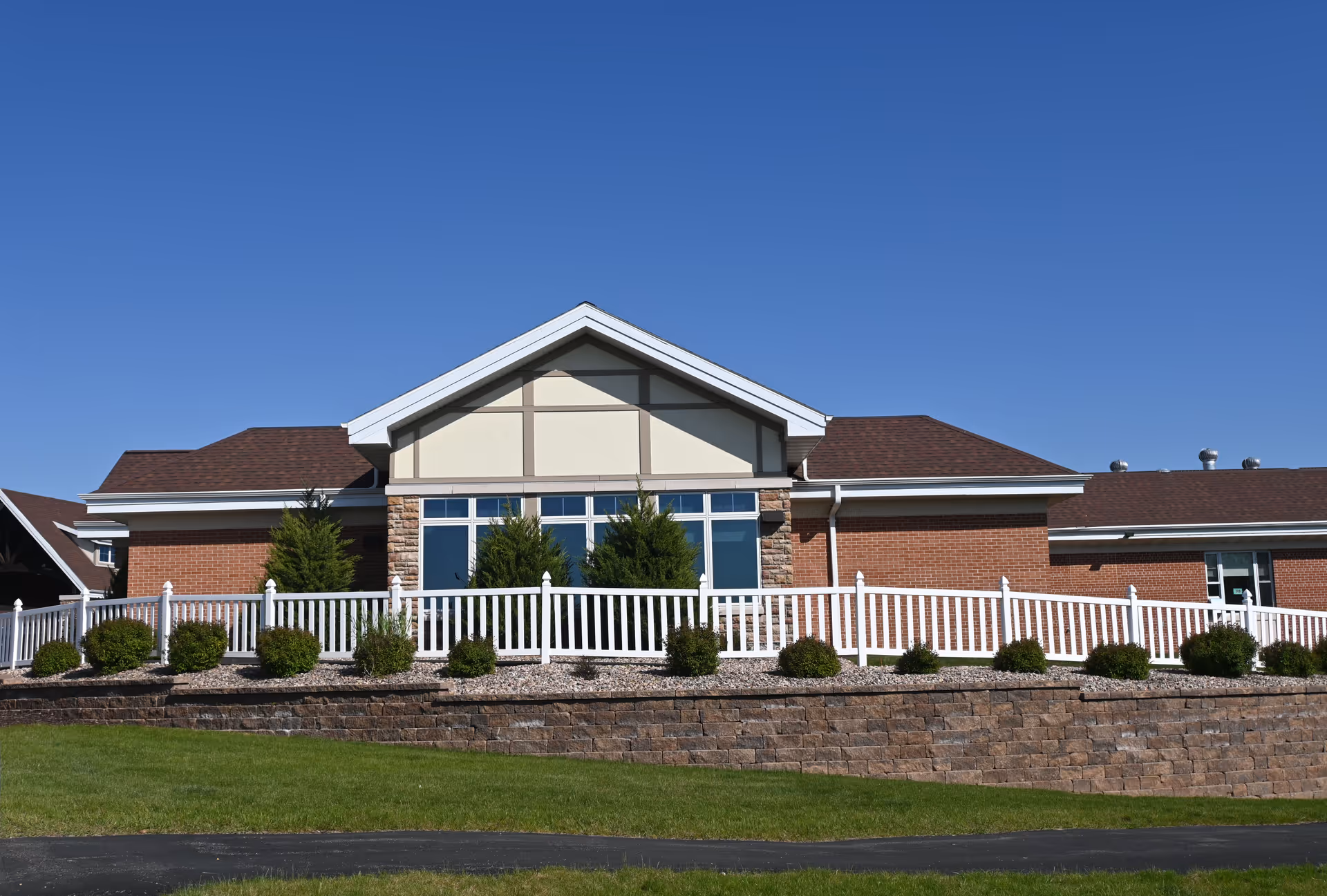 Exterior view of a single-story brick building with a gabled roof, large windows, white railing, and landscaped bushes in front under a clear blue sky.