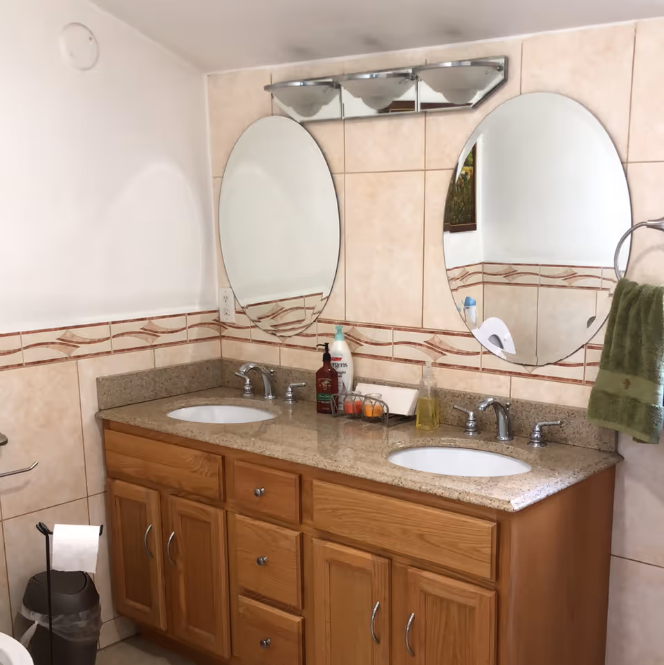 Bathroom with a double sink vanity featuring a granite countertop, wooden cabinets, two oval mirrors mounted on a tiled wall, a light fixture above the mirrors, a green towel hanging on a ring, and various toiletries on the countertop.