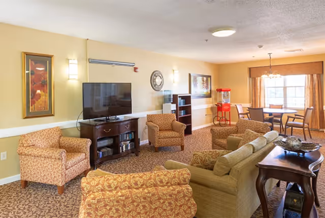 A cozy living room area in a senior living facility with patterned armchairs, a sofa, a wooden TV stand with a flat-screen television, a water dispenser, a popcorn machine, a dining table with chairs near a window with curtains, and decorative wall art.