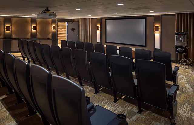 Interior view of a small theater room with multiple rows of black cushioned seats facing a large blank projection screen. The room has dim wall sconces for lighting, a popcorn machine on the right side, and a ceiling-mounted projector.