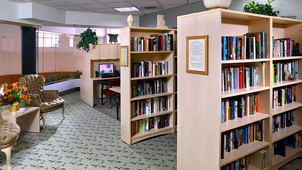 Interior view of a senior living facility library area with light wood bookshelves filled with books, a small computer desk with a monitor, upholstered armchairs, a small table with a flower arrangement, and carpeted flooring with a patterned design.