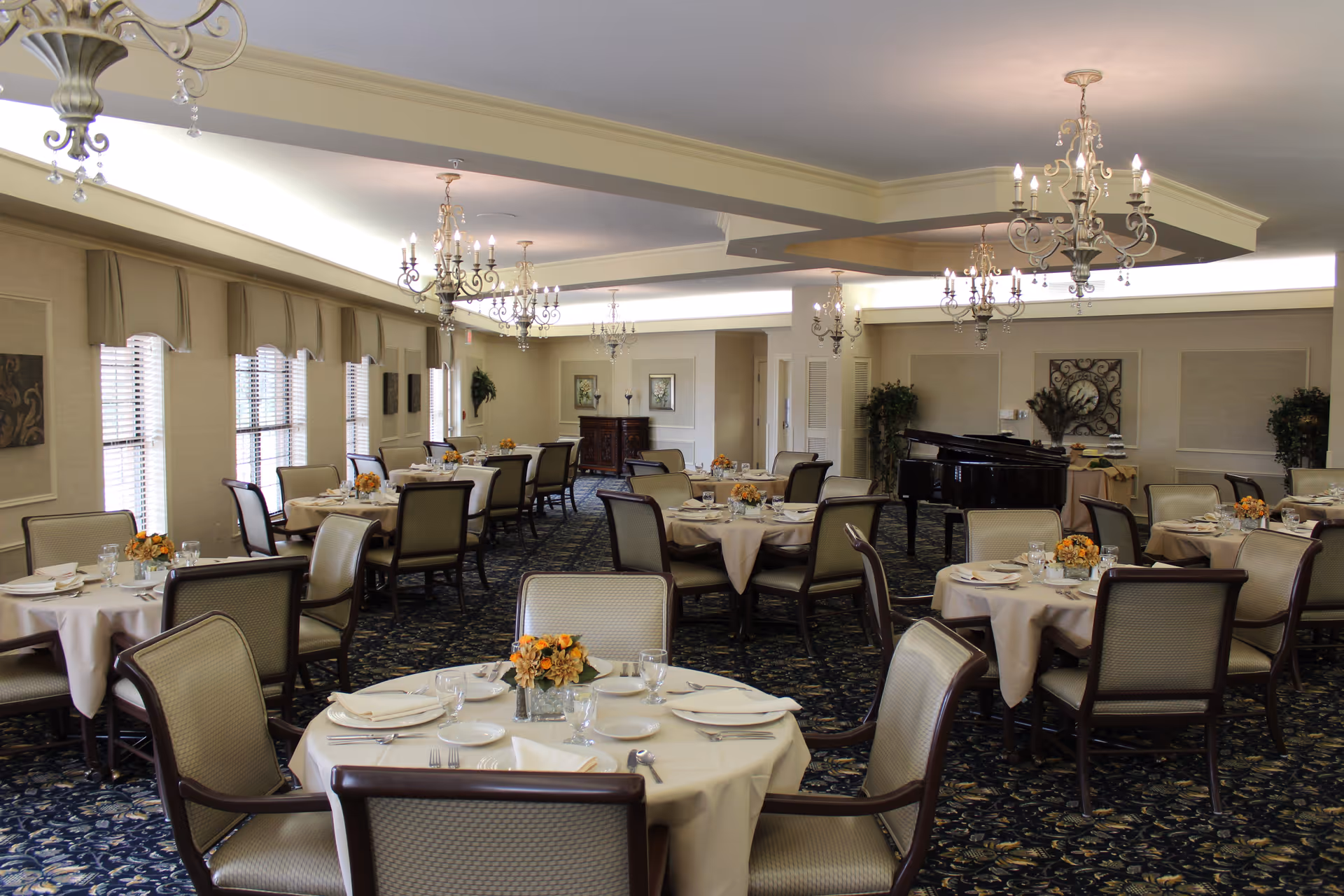 A spacious dining room with multiple round tables covered with white tablecloths, each set with plates, glasses, and silverware. The room features elegant chandeliers hanging from the ceiling, large windows with beige curtains, and a grand piano in the background. The carpet has a dark floral pattern, and there are decorative plants and framed artwork on the walls.