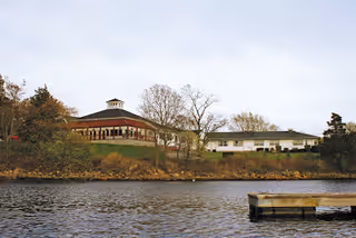 View across a body of water showing a building complex with a gazebo-like structure and a long single-story building surrounded by trees and shrubs under a cloudy sky.