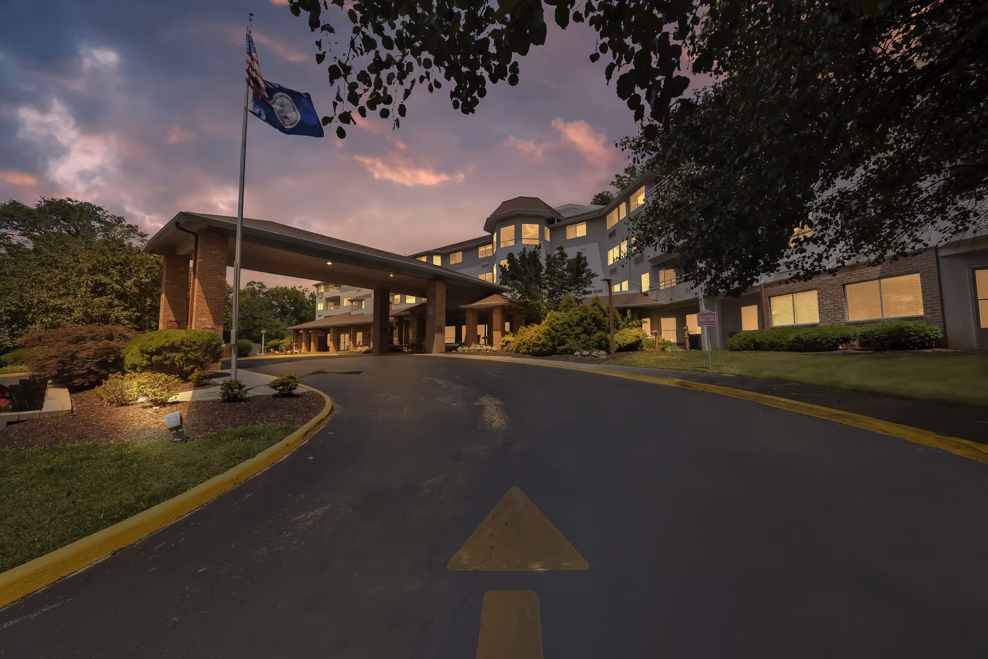 Exterior view of Holiday Elm Park Estates senior living facility at dusk, showing a curved driveway leading to a covered entrance with illuminated windows and surrounding greenery under a partly cloudy sky.