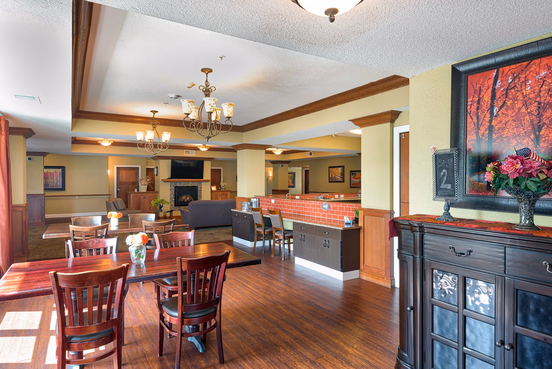 A spacious and well-lit common area in a senior living facility featuring wooden tables and chairs with flower vases, a cozy seating area with sofas near a fireplace and TV, warm lighting from chandeliers, and decorative artwork on the walls. The room has wooden flooring and a counter with bar stools.