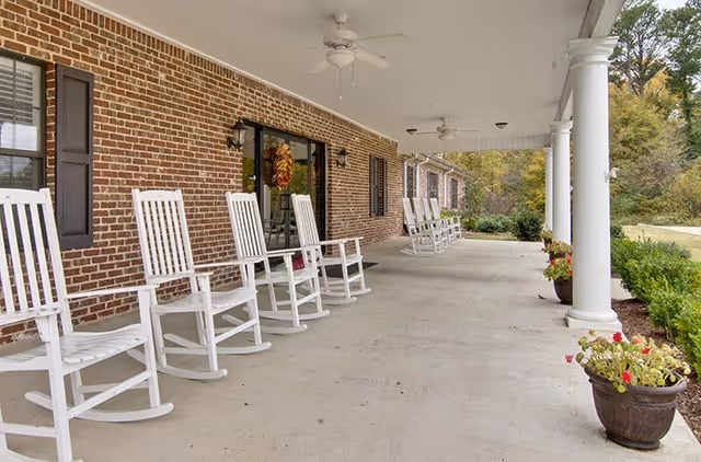 A covered outdoor porch area with white rocking chairs lined up against a brick wall. The porch has white columns, ceiling fans, and potted plants with flowers along the edge. Trees and greenery are visible in the background.