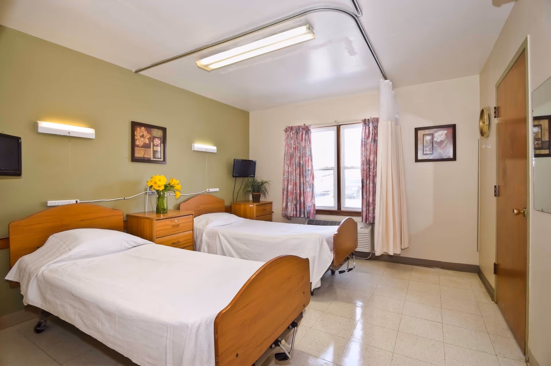 A simple, clean bedroom with two single beds with wooden headboards and white bedding. Between the beds is a wooden nightstand with a vase of yellow flowers. The walls are painted light green and beige, decorated with framed floral artwork. There are two wall-mounted lights above the beds, a small wall-mounted TV in the corner, a window with floral curtains, and a privacy curtain on a ceiling track. The floor is tiled, and there is a wooden door with a mirror on the right side.