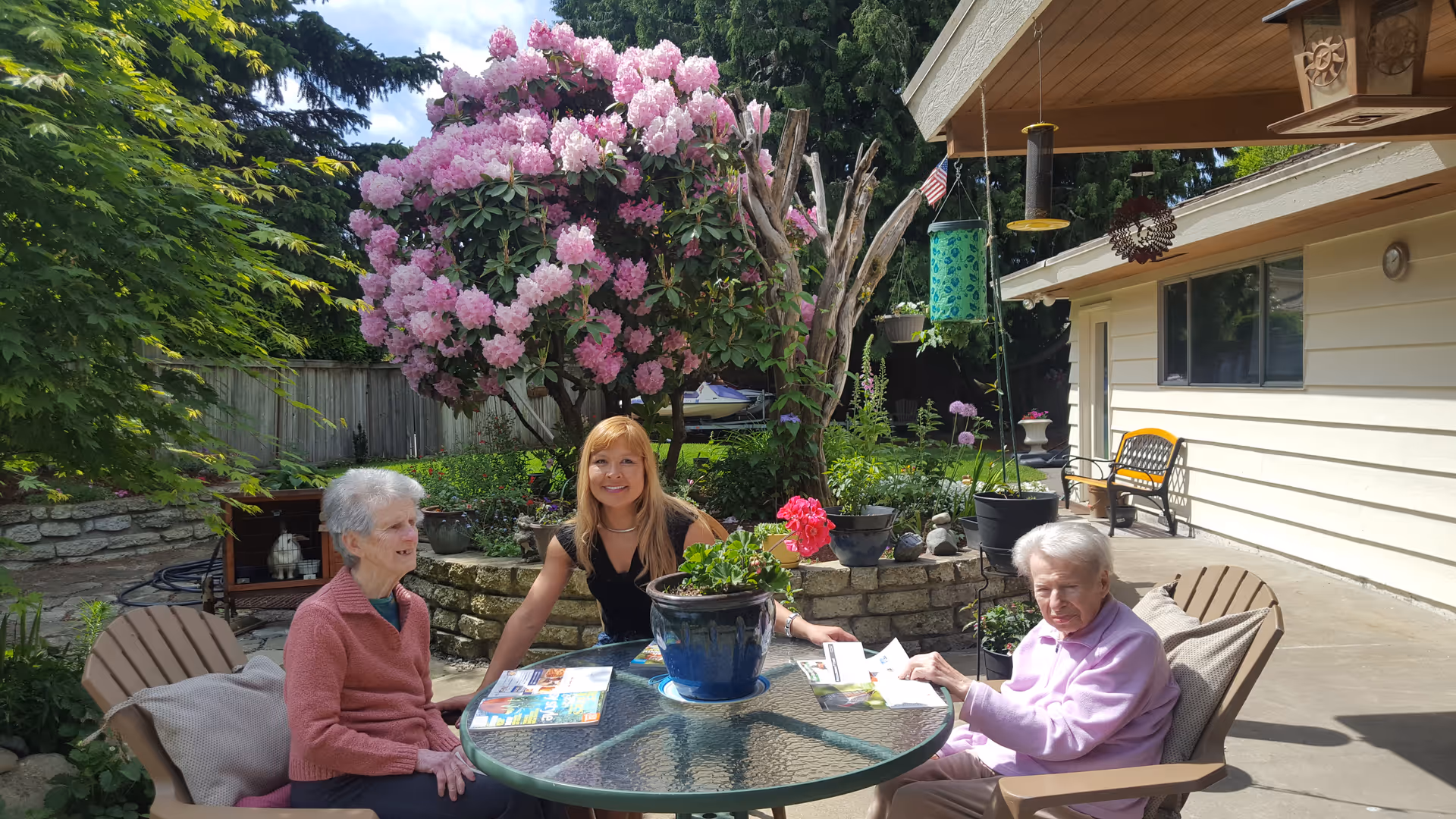 Three women sitting around a glass patio table with potted plants in a sunny backyard garden featuring a large pink-flowering bush.