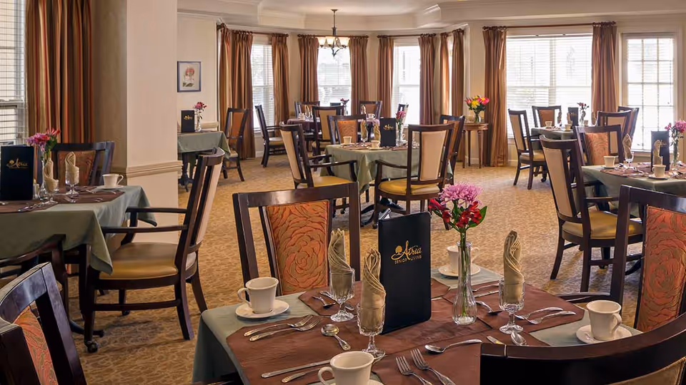 A well-lit dining room with multiple tables set for a meal. Each table has a green tablecloth with a brown placemat, neatly arranged silverware, cups, glasses with folded napkins, and a small vase with flowers. The chairs have wooden frames with cushioned seats and backs. Large windows with brown curtains allow natural light to fill the room.