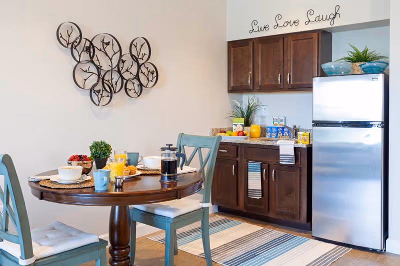 A cozy dining area with a round wooden table set for breakfast, featuring bowls, cups, a French press, and a small plant. Two blue wooden chairs with cushions are placed around the table. In the background, there is a kitchenette with dark wooden cabinets, a stainless steel refrigerator, and decorative plants on top. Above the cabinets, a wall decoration reads 'Live Love Laugh'. A striped rug lies on the floor beneath the table and chairs.