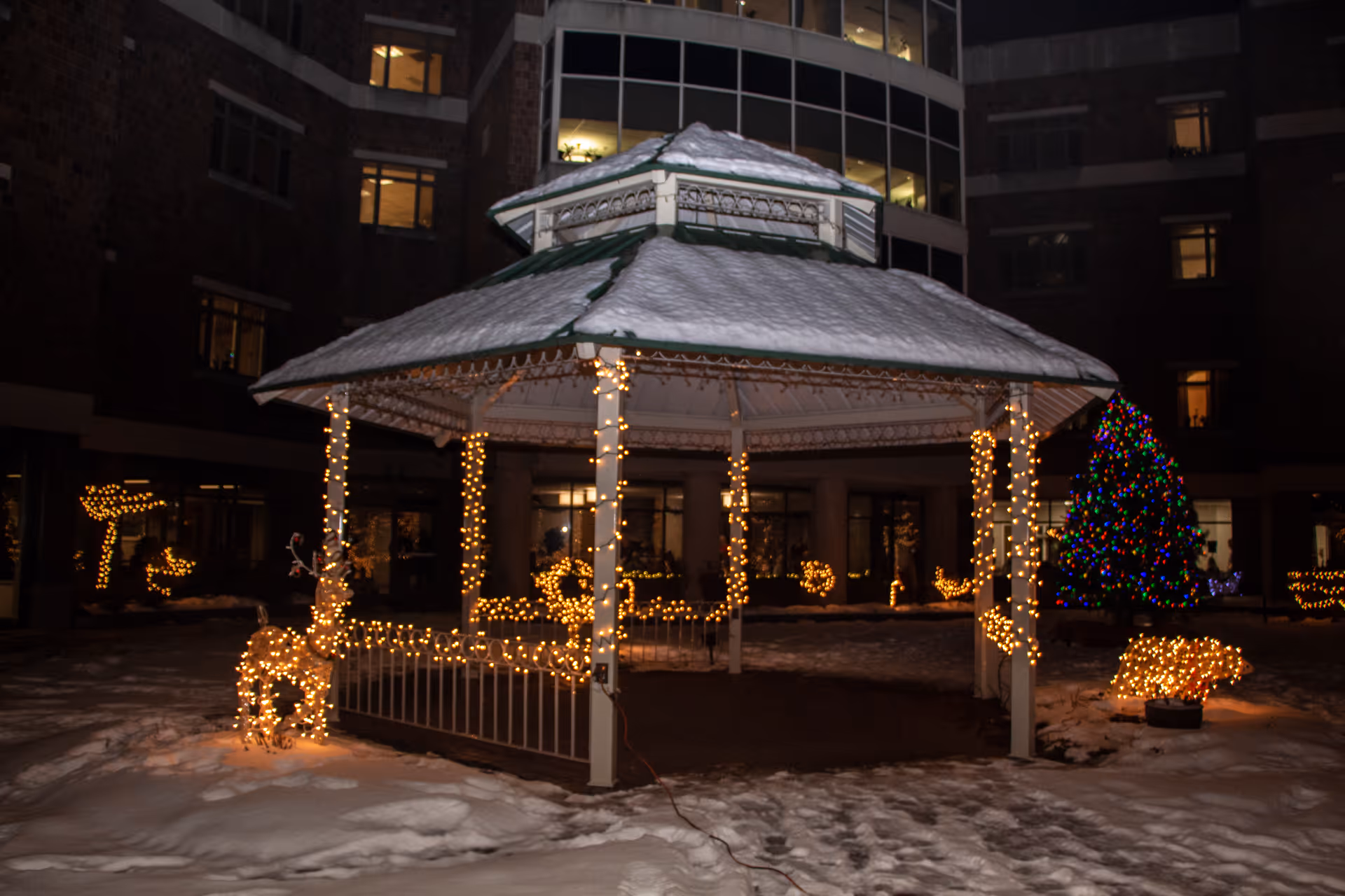 Snow-covered gazebo in a courtyard decorated with string lights and a lit Christmas tree at night.