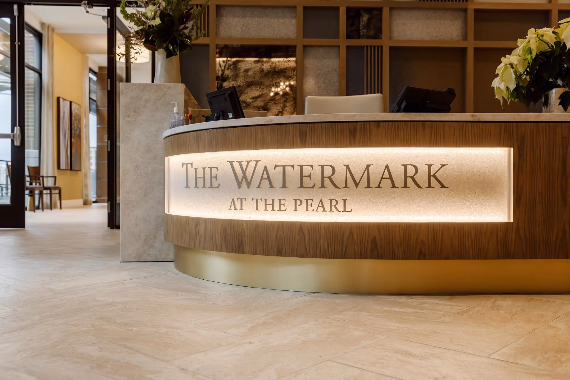 Reception desk area with a curved wooden front panel illuminated with the text 'The Watermark at the Pearl'. The background shows a modern interior with plants and a hallway leading to other rooms.