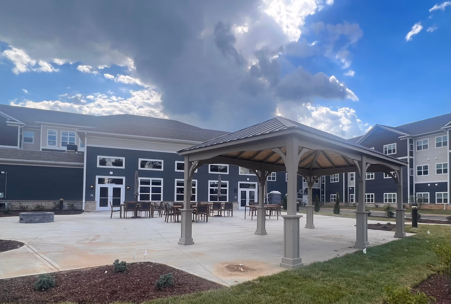 Outdoor courtyard with a covered pavilion, patio tables, and a multi-story building under a cloudy sky.