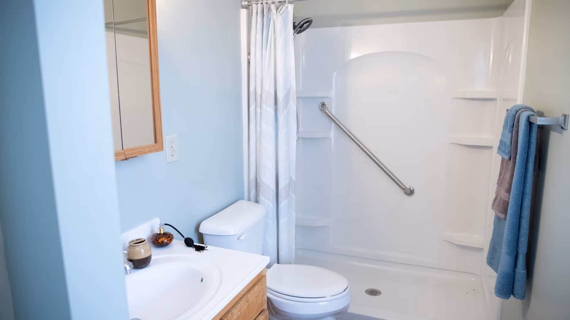 A clean bathroom with light blue walls featuring a white sink with wooden cabinet, a toilet, and a white shower with a grab bar and a shower curtain. Towels are hanging on a rack on the right wall.