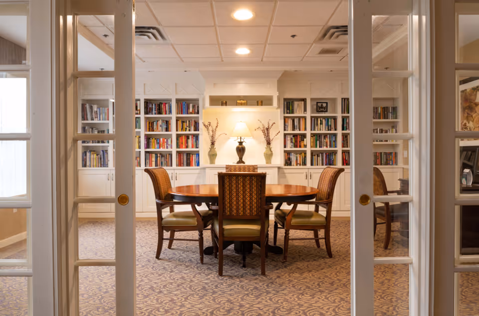 A cozy library room with built-in white bookshelves filled with books, a round wooden table surrounded by four upholstered chairs, and a table lamp with two vases on either side, viewed through glass-paneled double doors.