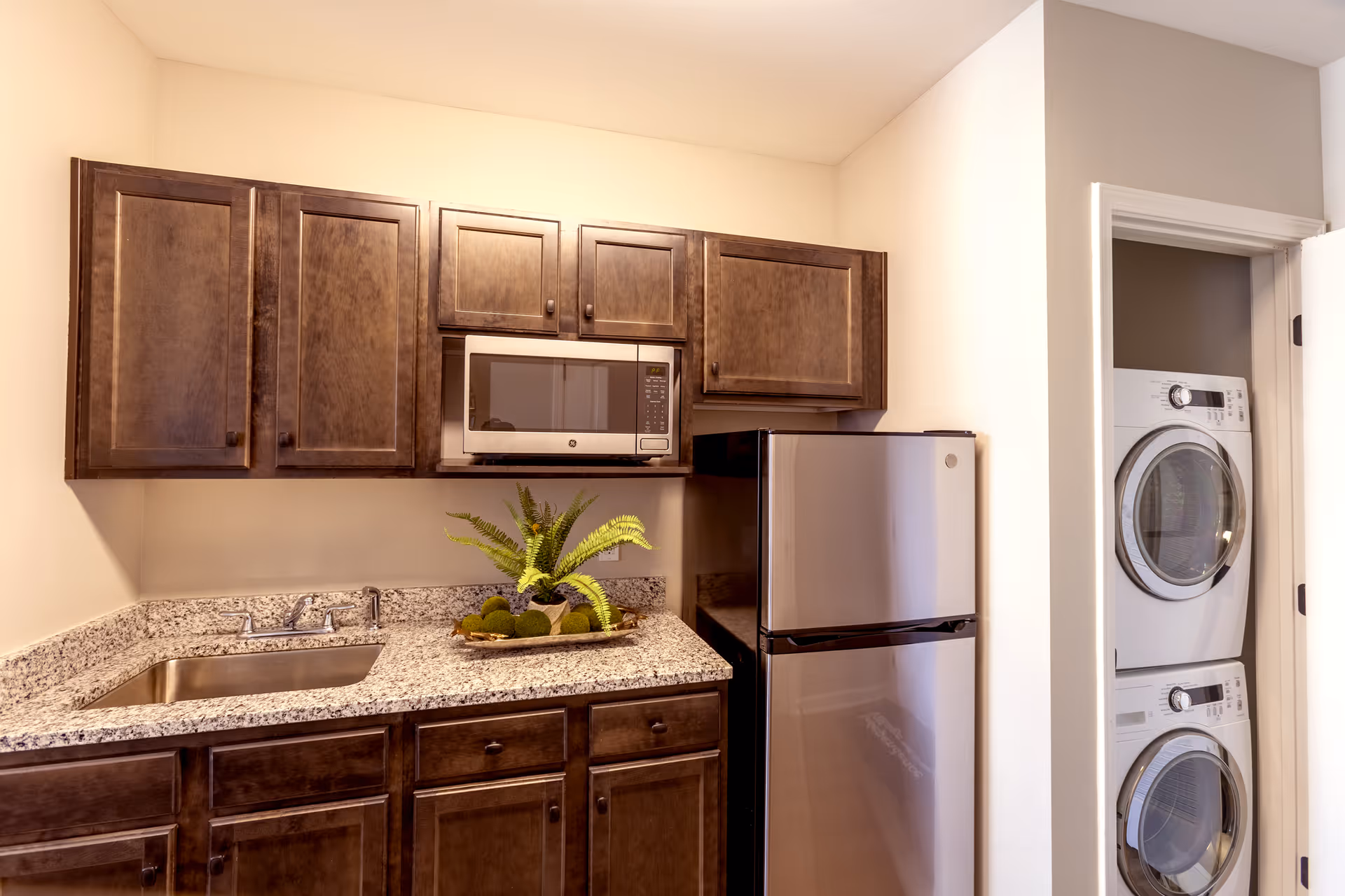 A compact kitchen area with dark wooden cabinets, a granite countertop, a stainless steel sink, a microwave oven, and a stainless steel refrigerator. To the right, there is a small laundry closet with a stacked washer and dryer visible through an open door.