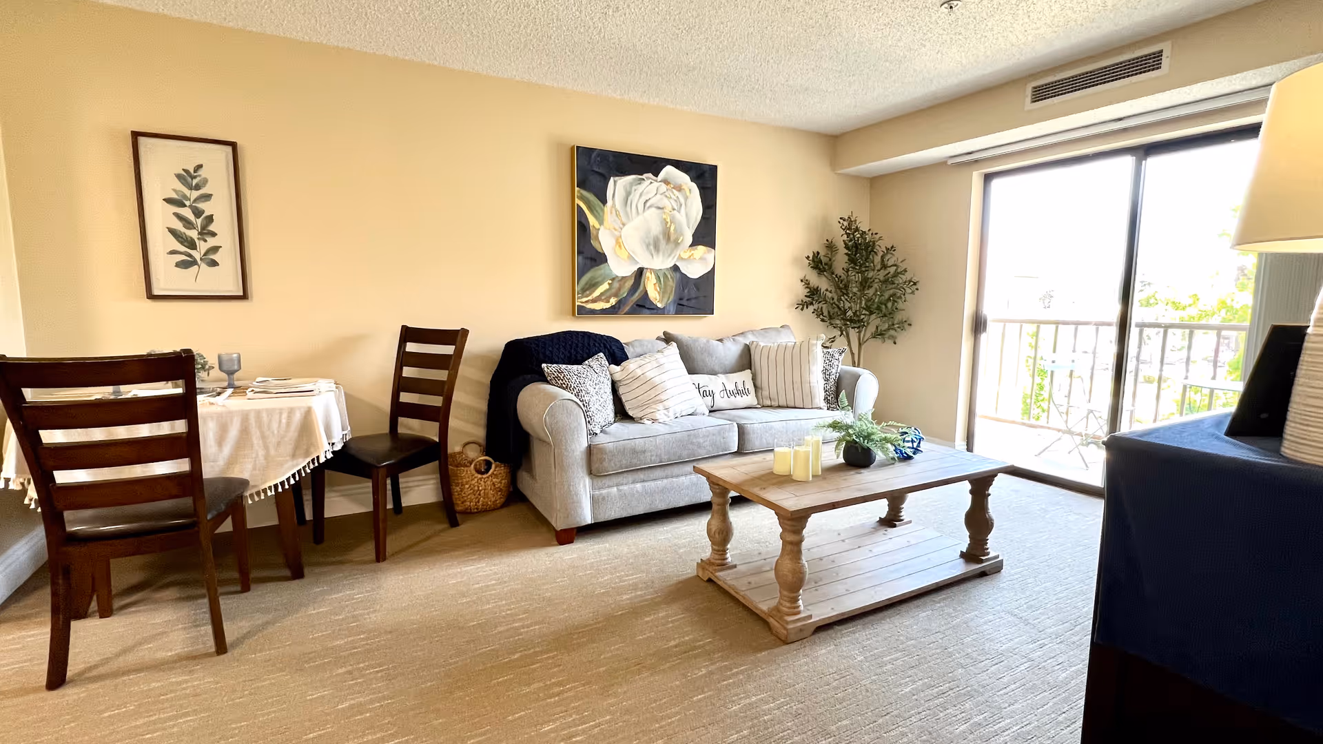Bright living room with a sofa, coffee table, small dining table with chairs, wall art, and a sliding glass door to a balcony.