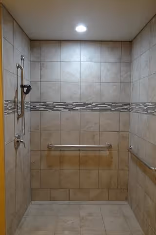 A tiled shower area with beige and light brown tiles, featuring a horizontal decorative strip of smaller rectangular tiles in the middle. The shower has a metal grab bar on the back wall and another on the right wall, along with a handheld showerhead mounted on the left wall. The ceiling has a recessed light.