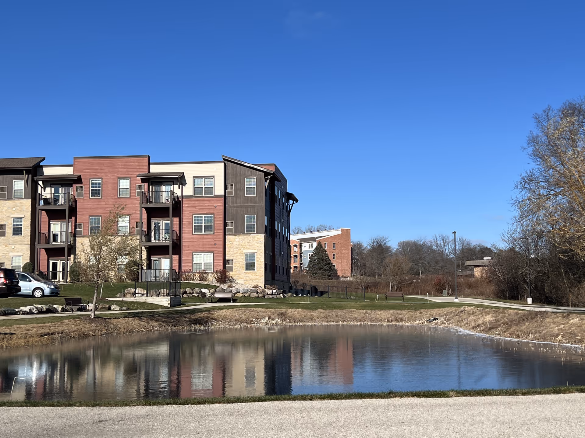 Exterior view of a multi-story senior living facility building with balconies, situated next to a small pond with clear reflections of the building and trees. The sky is clear and blue, and there is a paved path and some parked cars visible near the building.