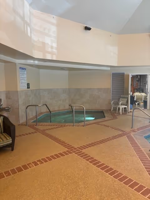 Indoor pool area with a small rectangular pool surrounded by beige and brick-patterned flooring. There are metal handrails leading into the pool, a chair with striped cushions on the left, and two white plastic chairs near an open door on the right. The walls are tiled halfway up and painted beige above, with a high ceiling and some equipment visible through the open door.