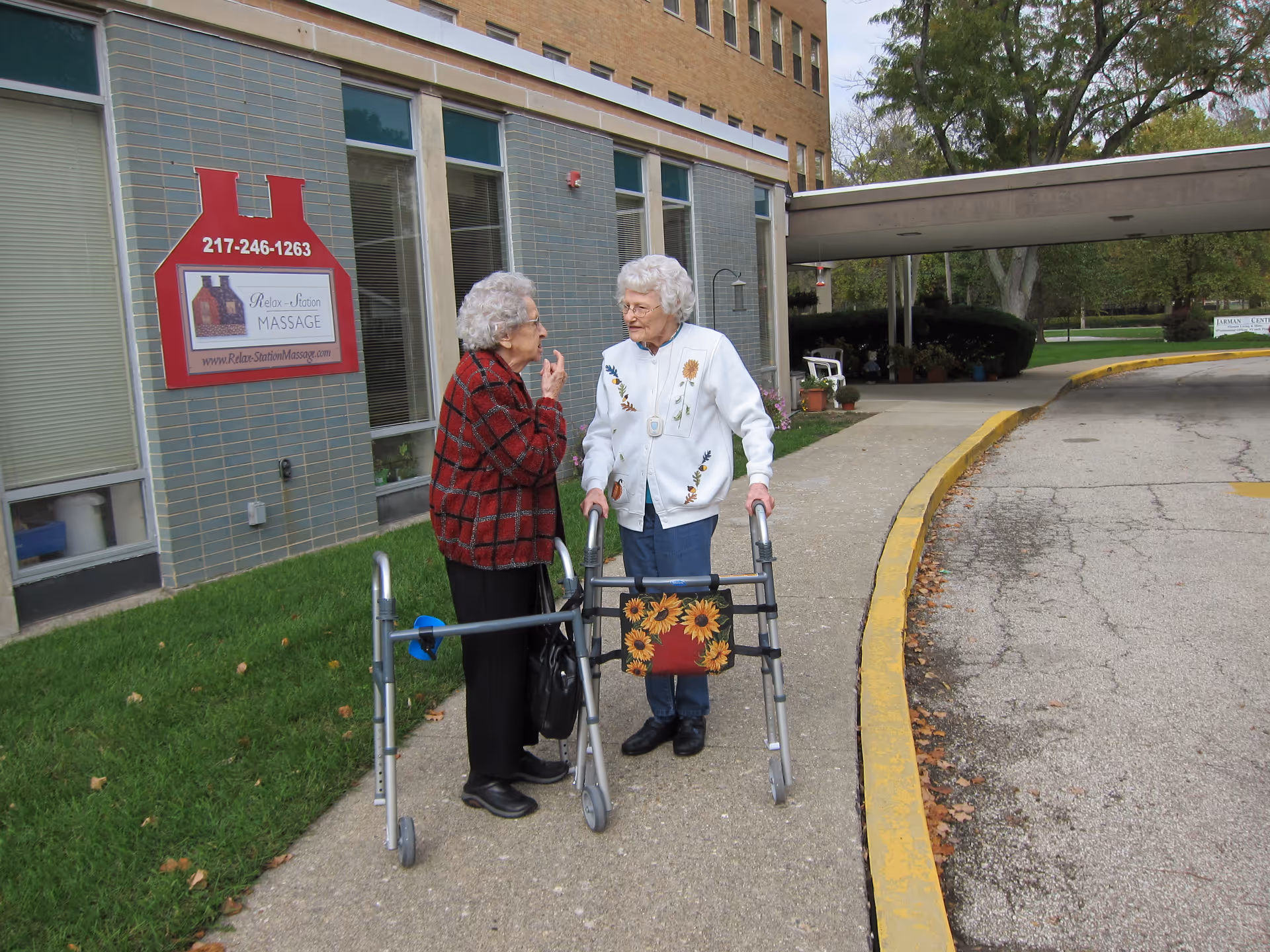 Two elderly women standing outside near a building entrance, each using a walker. One woman is wearing a red plaid jacket and the other is wearing a white jacket with floral embroidery. They appear to be engaged in conversation. There is a sign on the building advertising Relax-Station Massage with a phone number and website. The area has a sidewalk, grass, and a driveway with a yellow curb.